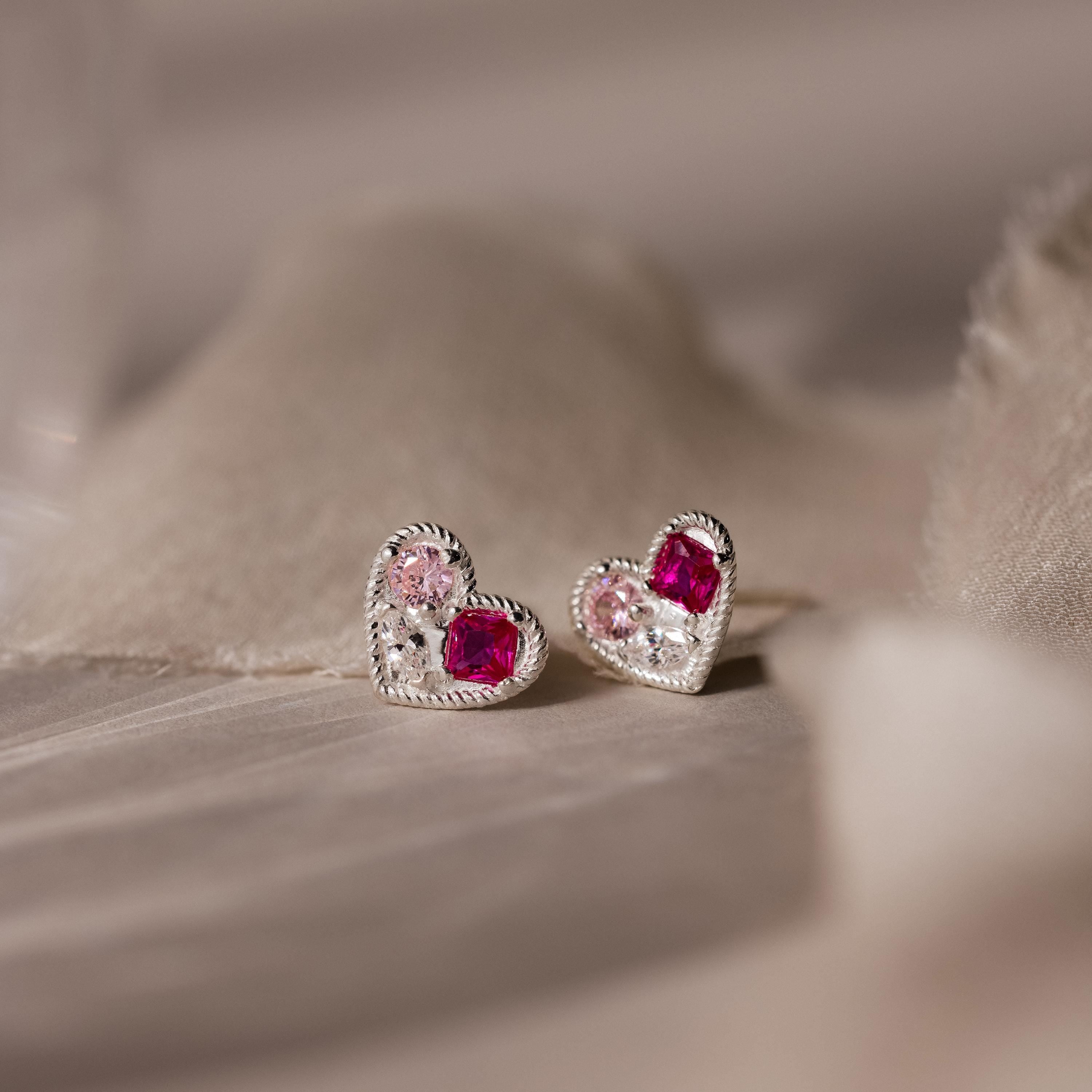 Mixed Birthstone Heart Studs feature silver heart-shaped earrings with customizable pink and red gemstones, beautifully displayed on soft beige fabric.
