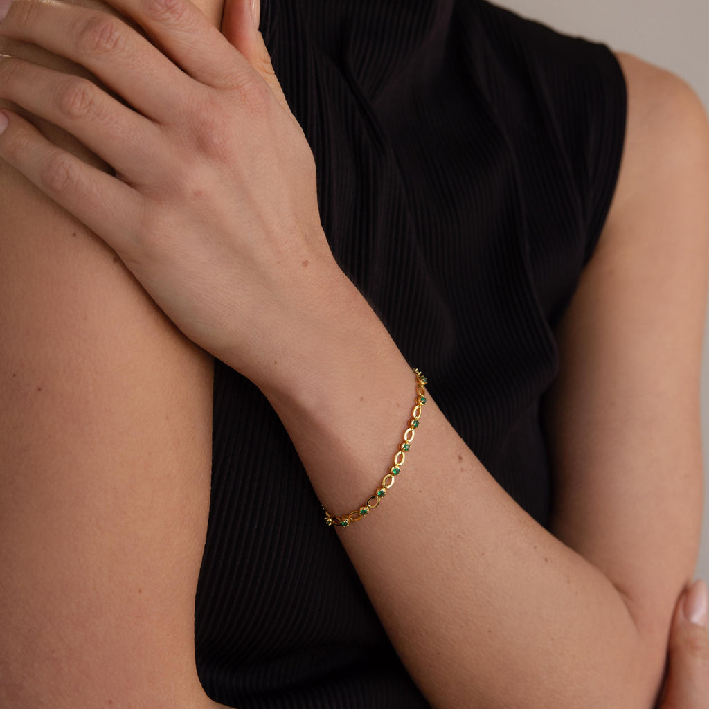 A person wears a sleeveless black top and an Emerald Chain Tennis Bracelet—a delicate gold bracelet adorned with emerald stones—on their wrist.