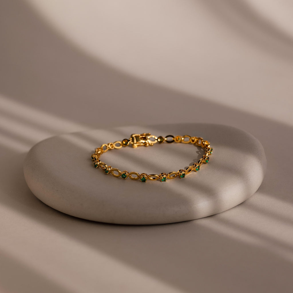 The Emerald Chain Tennis Bracelet features vibrant emerald stones set in gold, elegantly displayed on a smooth round stone under soft natural light.