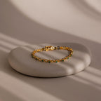 The Emerald Chain Tennis Bracelet features vibrant emerald stones set in gold, elegantly displayed on a smooth round stone under soft natural light.