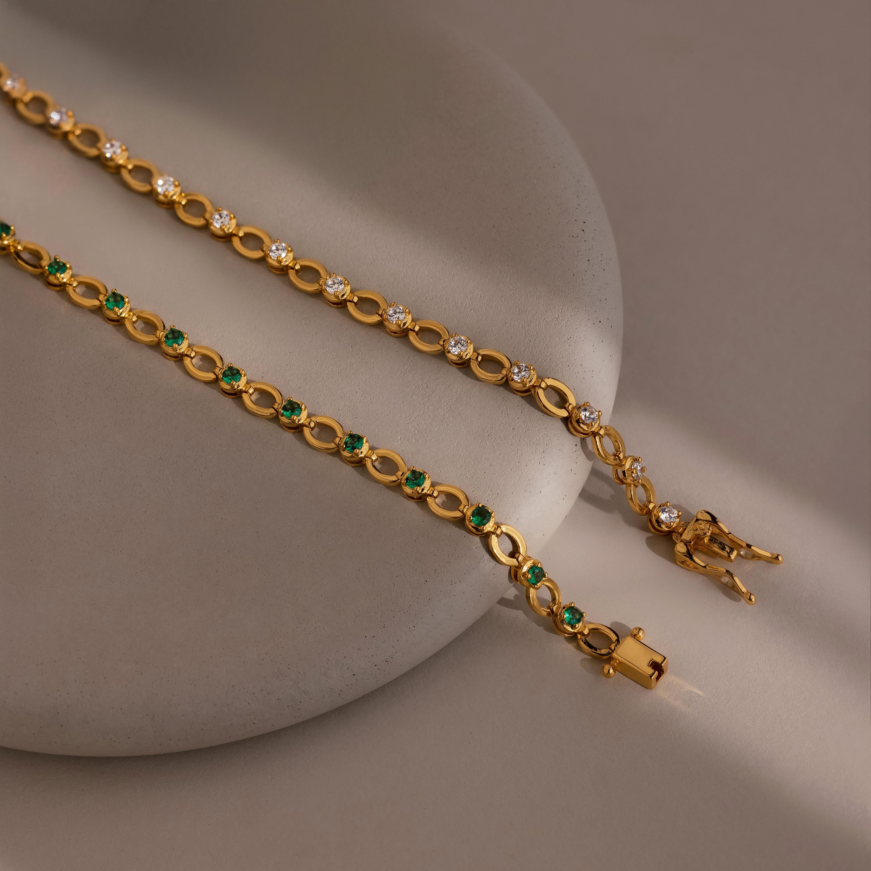 The Emerald Chain Tennis Bracelet featuring emerald stones, along with a white stone chain bracelet, are displayed on a round beige surface.