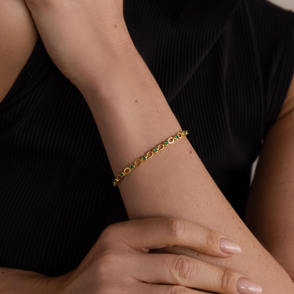 Close-up of a woman's arm wearing the Emerald Chain Tennis Bracelet, featuring delicate gold with small emerald stones, set against a black top.