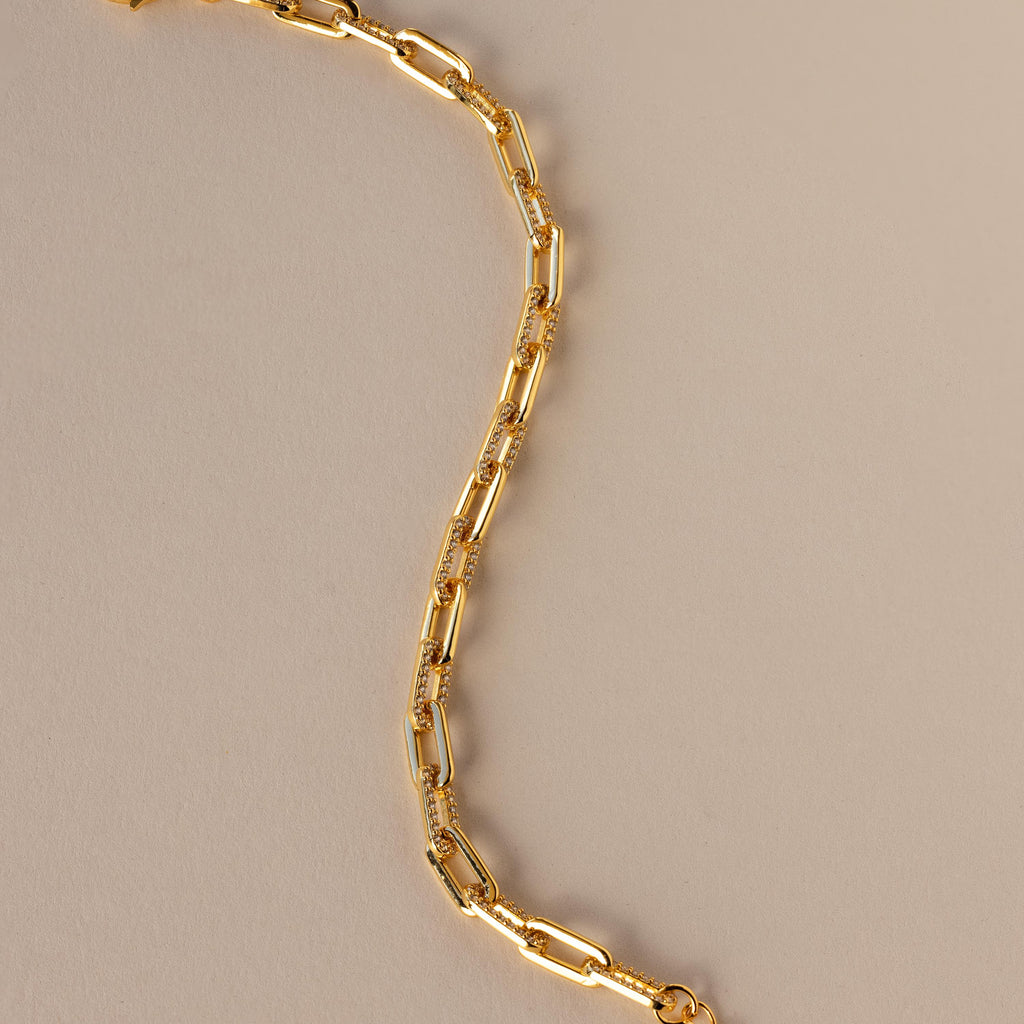 The Pave Paperclip Chain Bracelet features gold rectangular links with sparkling accents, shown on a beige background.