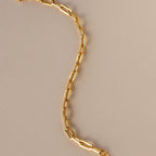 The Pave Paperclip Chain Bracelet features gold rectangular links with sparkling accents, shown on a beige background.