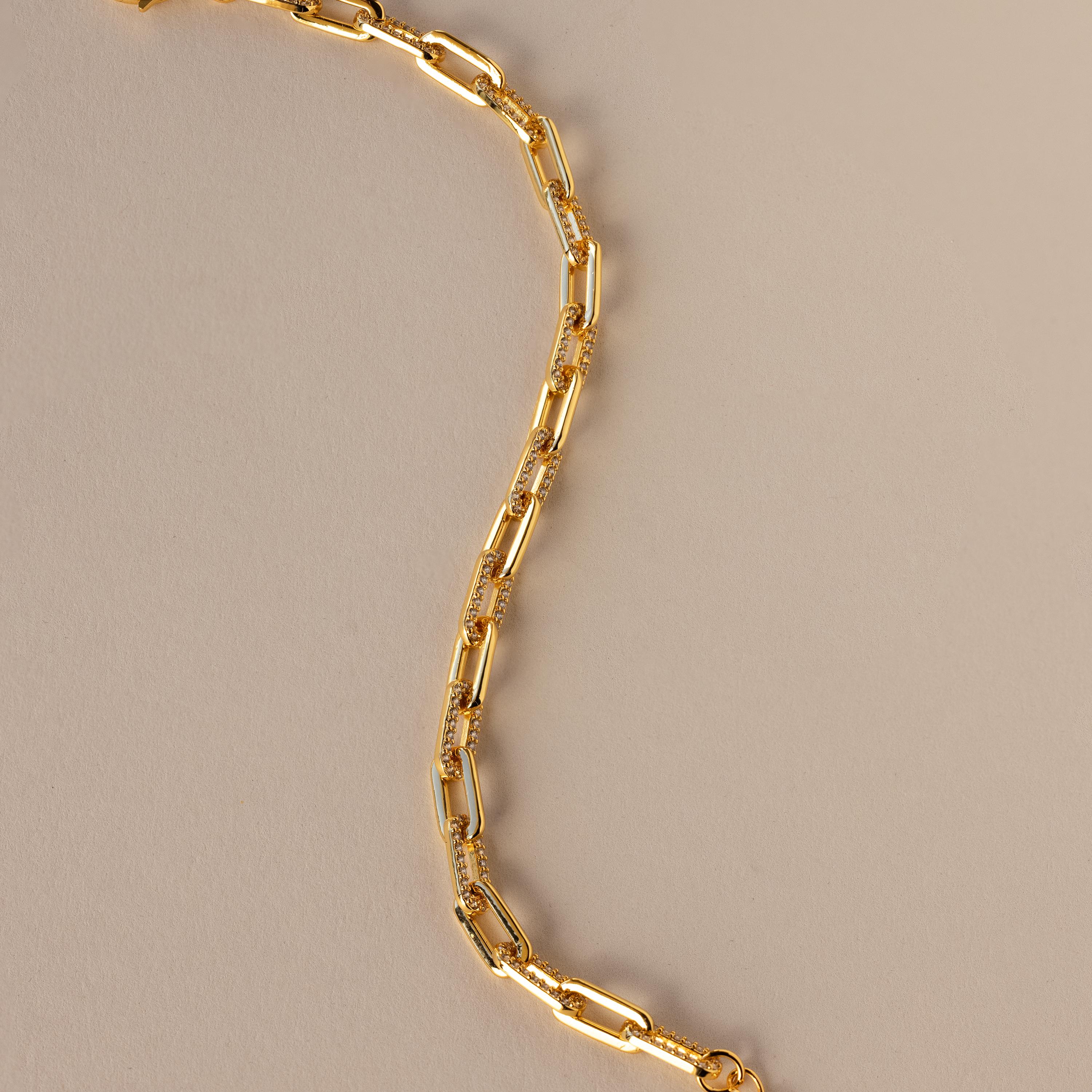 The Pave Paperclip Chain Bracelet features gold rectangular links with sparkling accents, shown on a beige background.