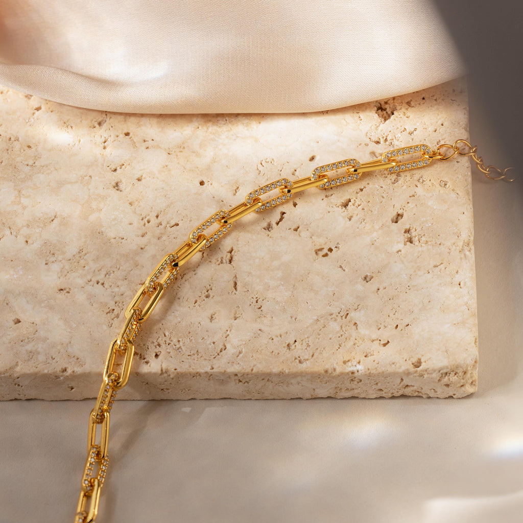 The Pave Paperclip Chain Bracelet features rectangular links and is displayed on a textured beige stone surface with soft lighting, highlighting its refined and elegant allure.