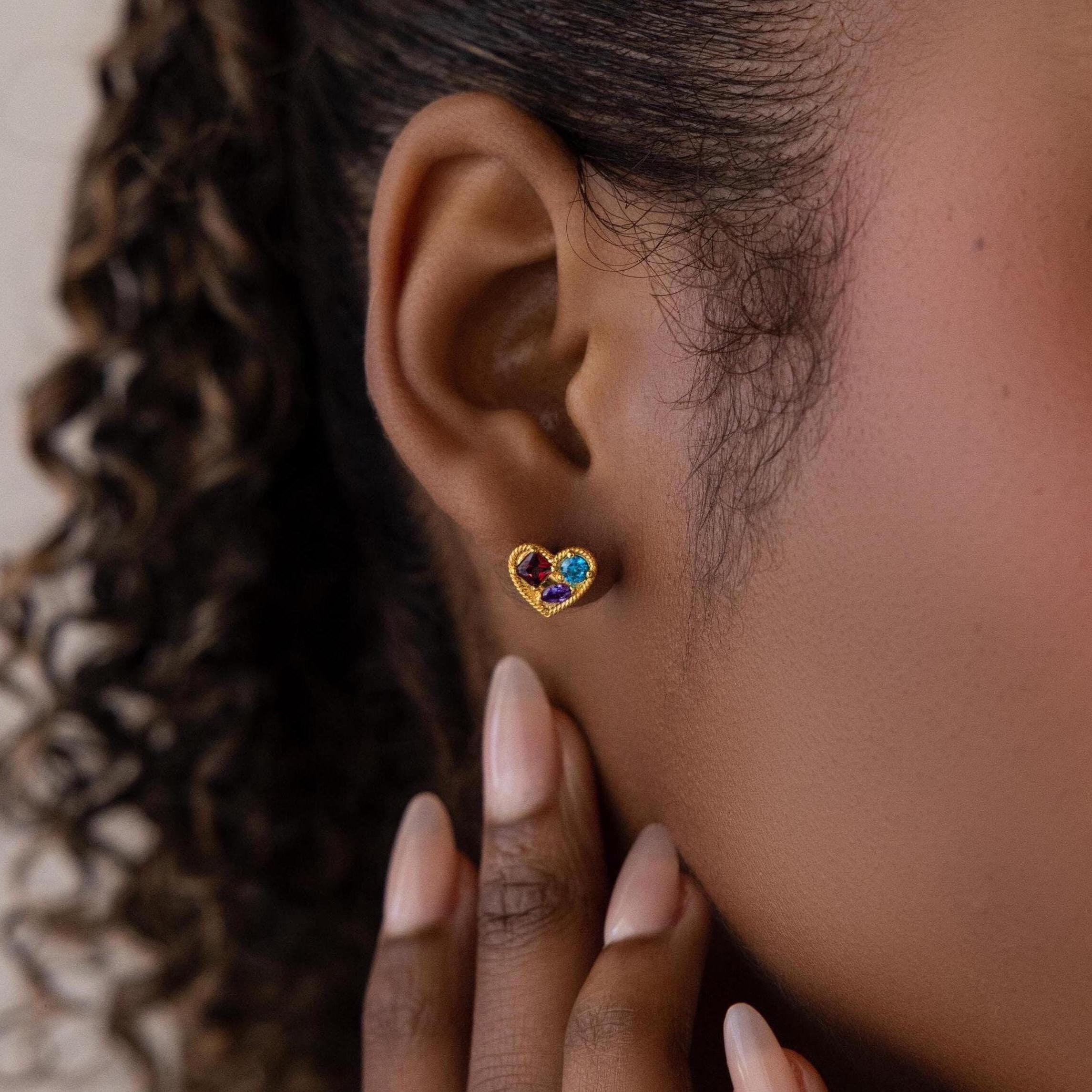 Close-up of a person wearing Mixed Birthstone Heart Studs—gold heart-shaped earrings featuring customizable red, blue, and green gemstones.