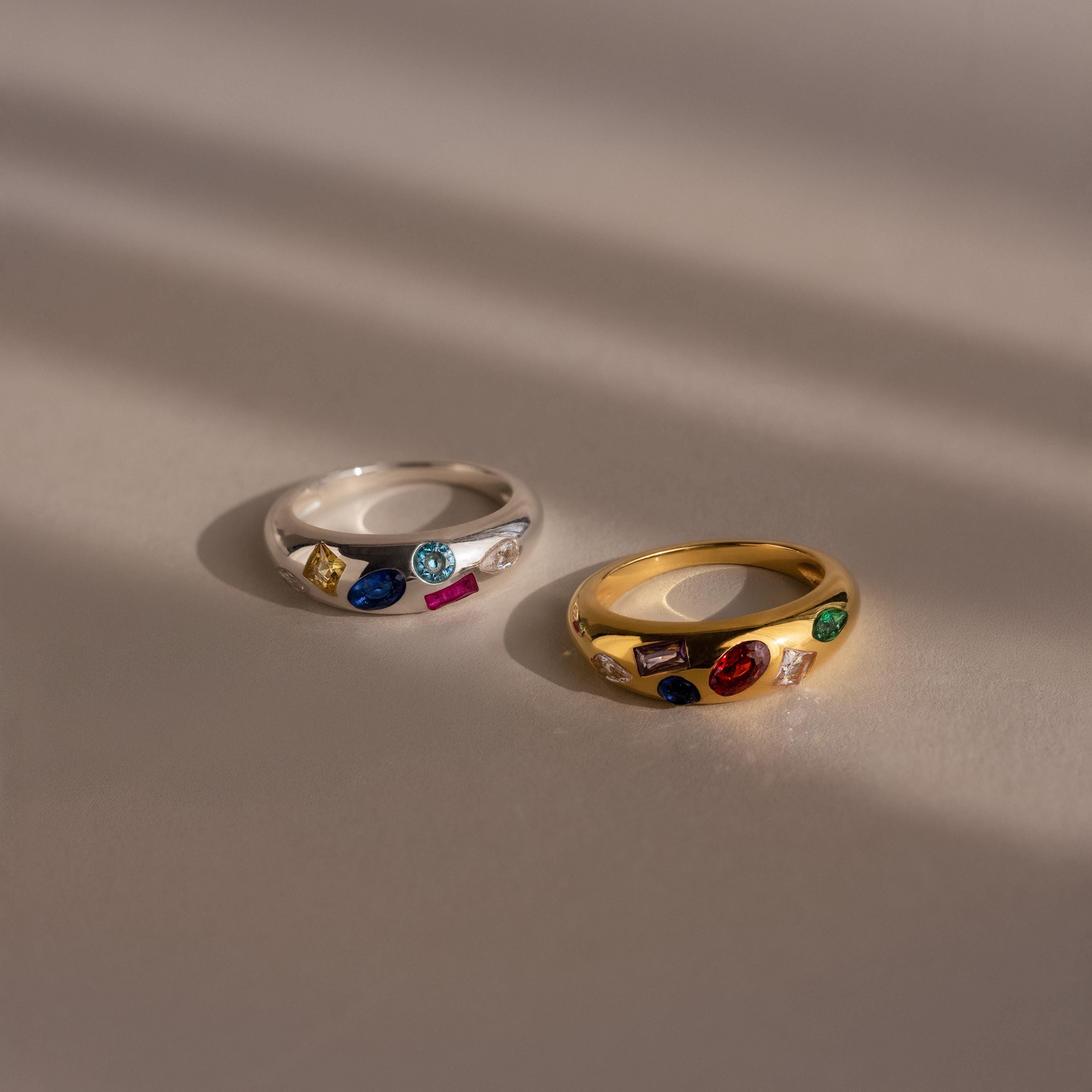 The Mixed Birthstone Dome Ring shines in both silver and gold on a smooth surface, highlighted by soft light and shadows—a stunning showcase of customizable elegance.