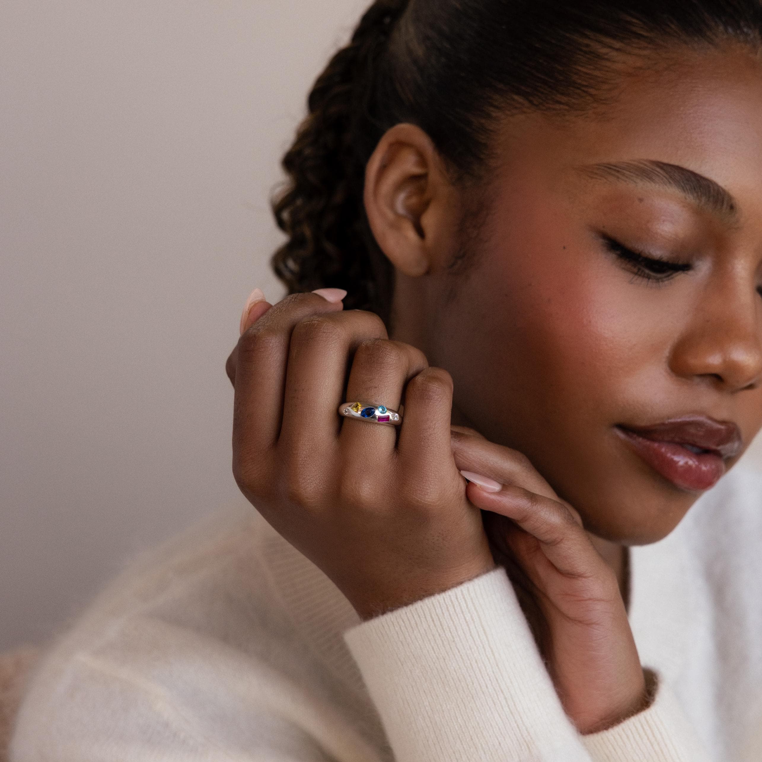 A woman with closed eyes gently touches her face, displaying the elegant Mixed Birthstone Dome Ring on her finger.