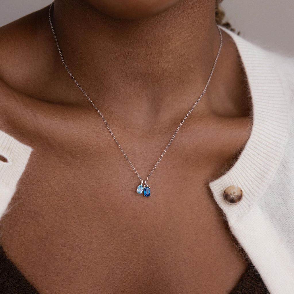 Model wears silver cable chain with aquamarine pear birthstone charm and blue zircon baguette birthstone charm