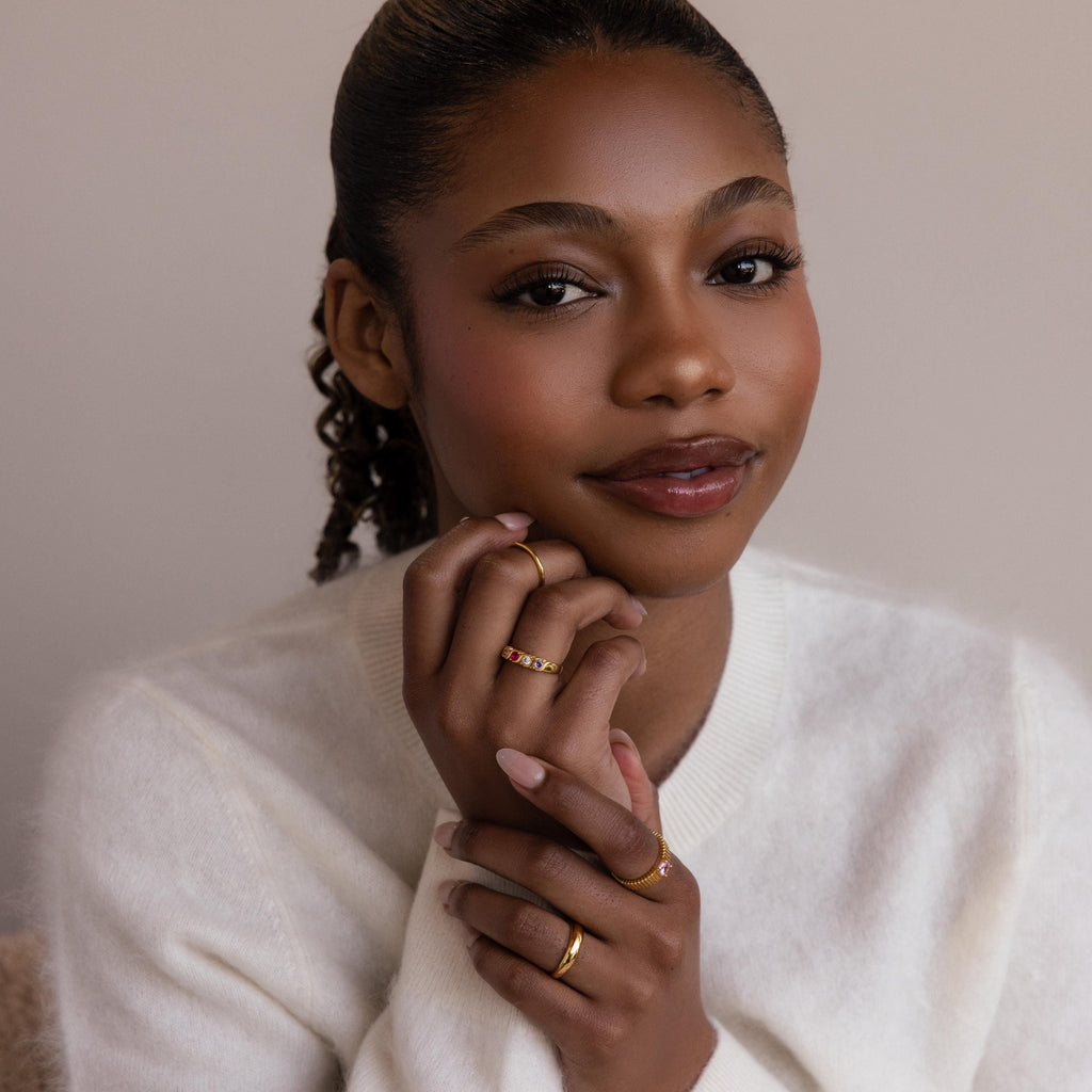 A woman with natural makeup and gold rings, including a delicate Vintage Multiple Birthstone Ring, poses in a white sweater against a neutral background.