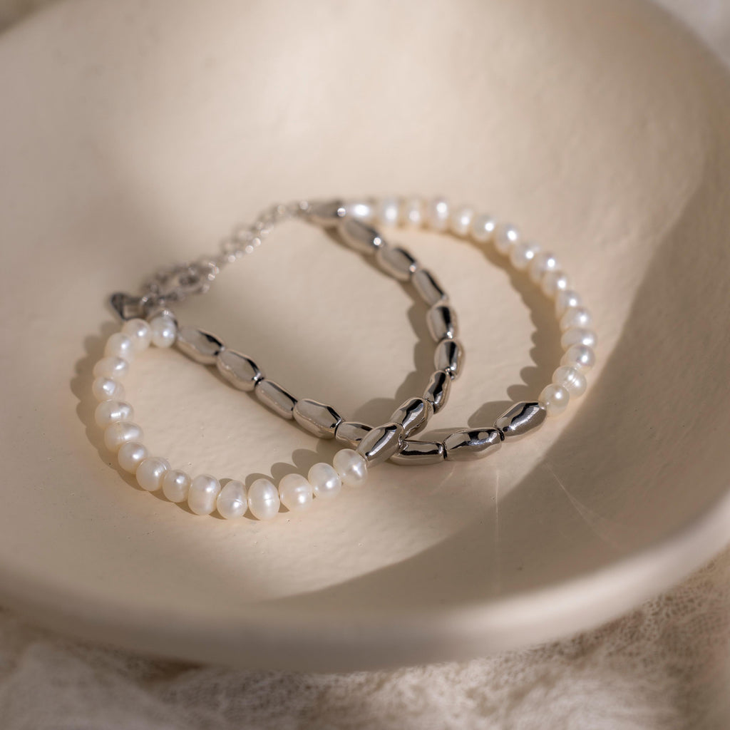 The Duo Beaded Pearl Bracelet, featuring delicate white pearls and silver beads, is elegantly displayed in a cream-colored dish.