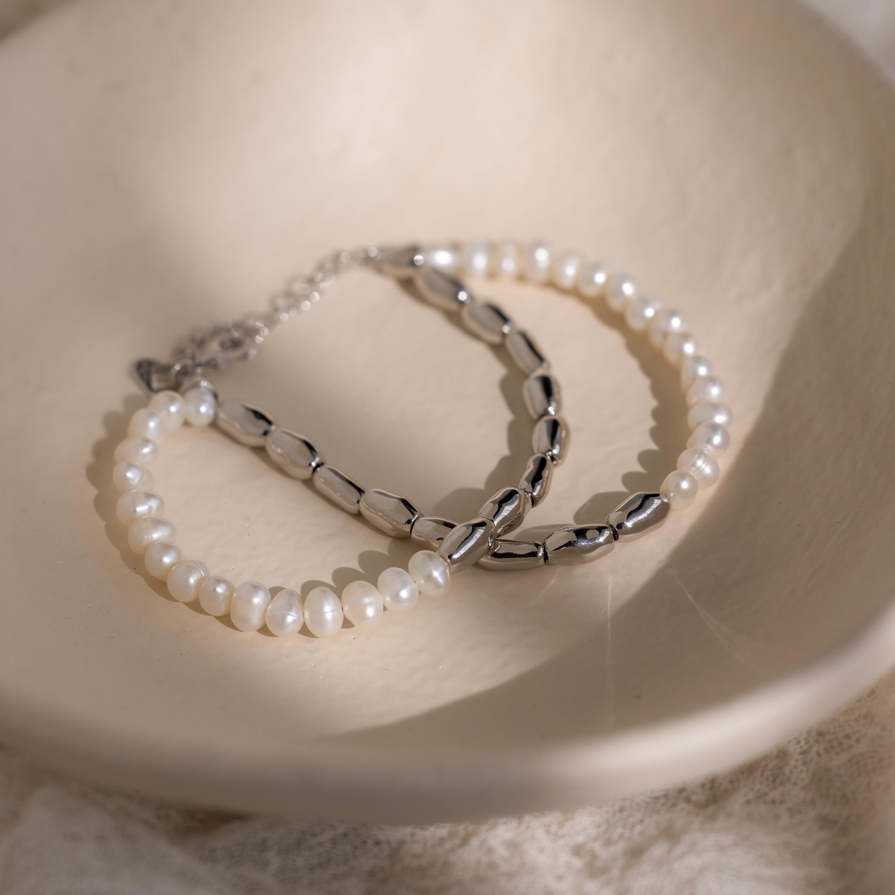 The Duo Beaded Pearl Bracelet, featuring delicate white pearls and silver beads, is elegantly displayed in a cream-colored dish.