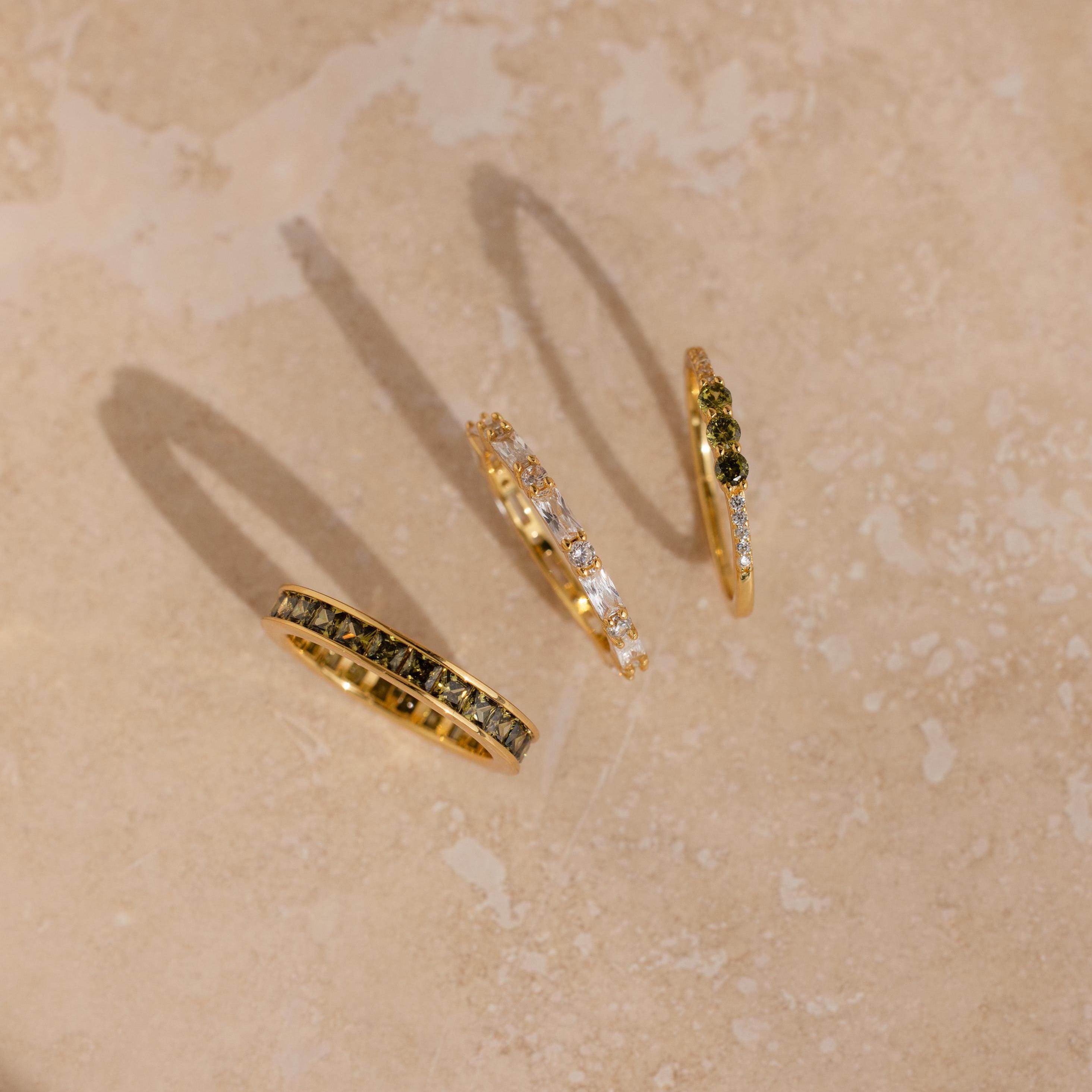 Overhead view of three gold peridot and diamond rings casting shadows on a beige surface.