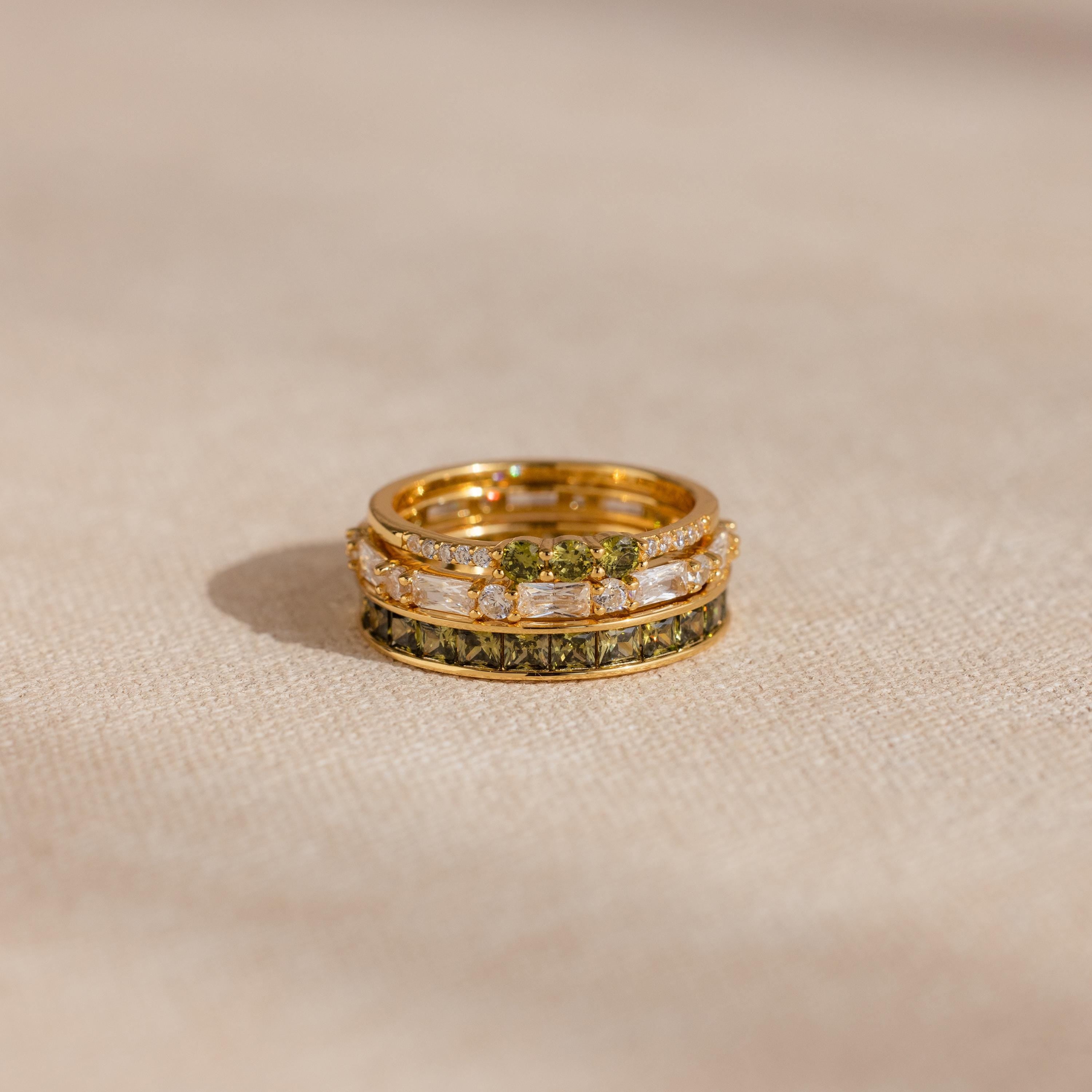 Close-up of three stacked gold peridot and diamond rings placed on a neutral fabric surface.