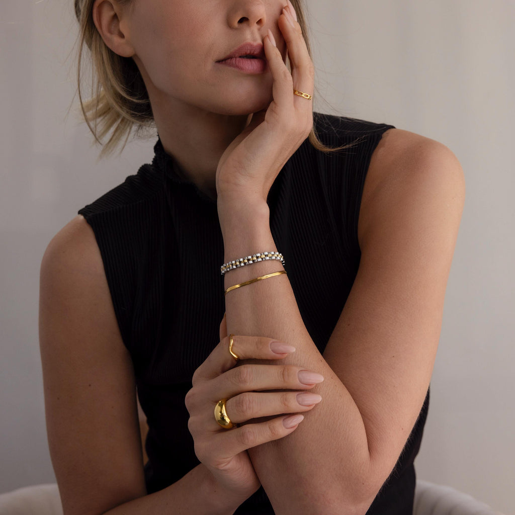 Woman in a sleeveless black top wears gold rings, bracelets, a beaded bracelet, and the Mixed Metal Link Bracelet, hand by her face.