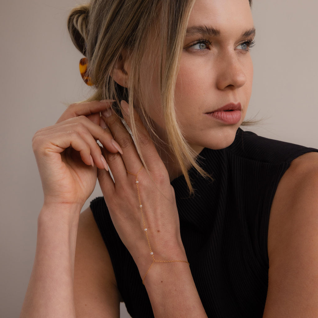 Wearing the Pearl Hand Chain Bracelet with delicate gold jewelry and a black sleeveless top, she gazes to the side, her hand near her face—a perfect touch of summer elegance.