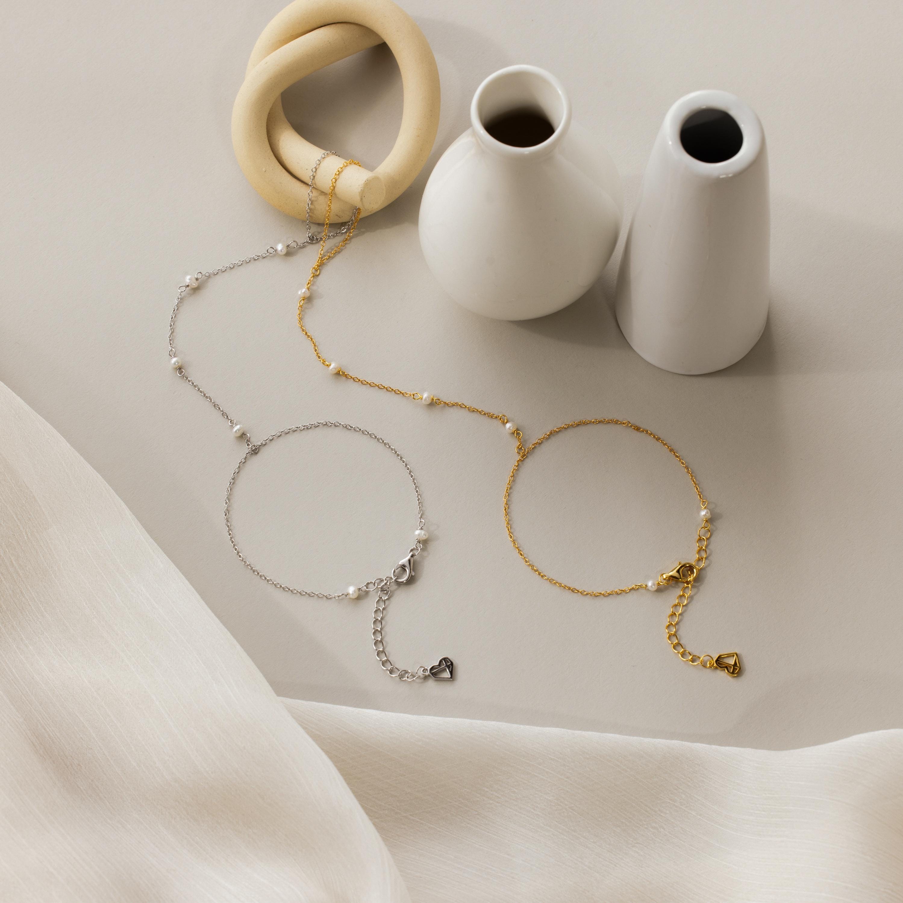 The Pearl Hand Chain Bracelet, shown near white vases on a light fabric surface, adds summer elegance to your style with its delicate gold and silver chain design.