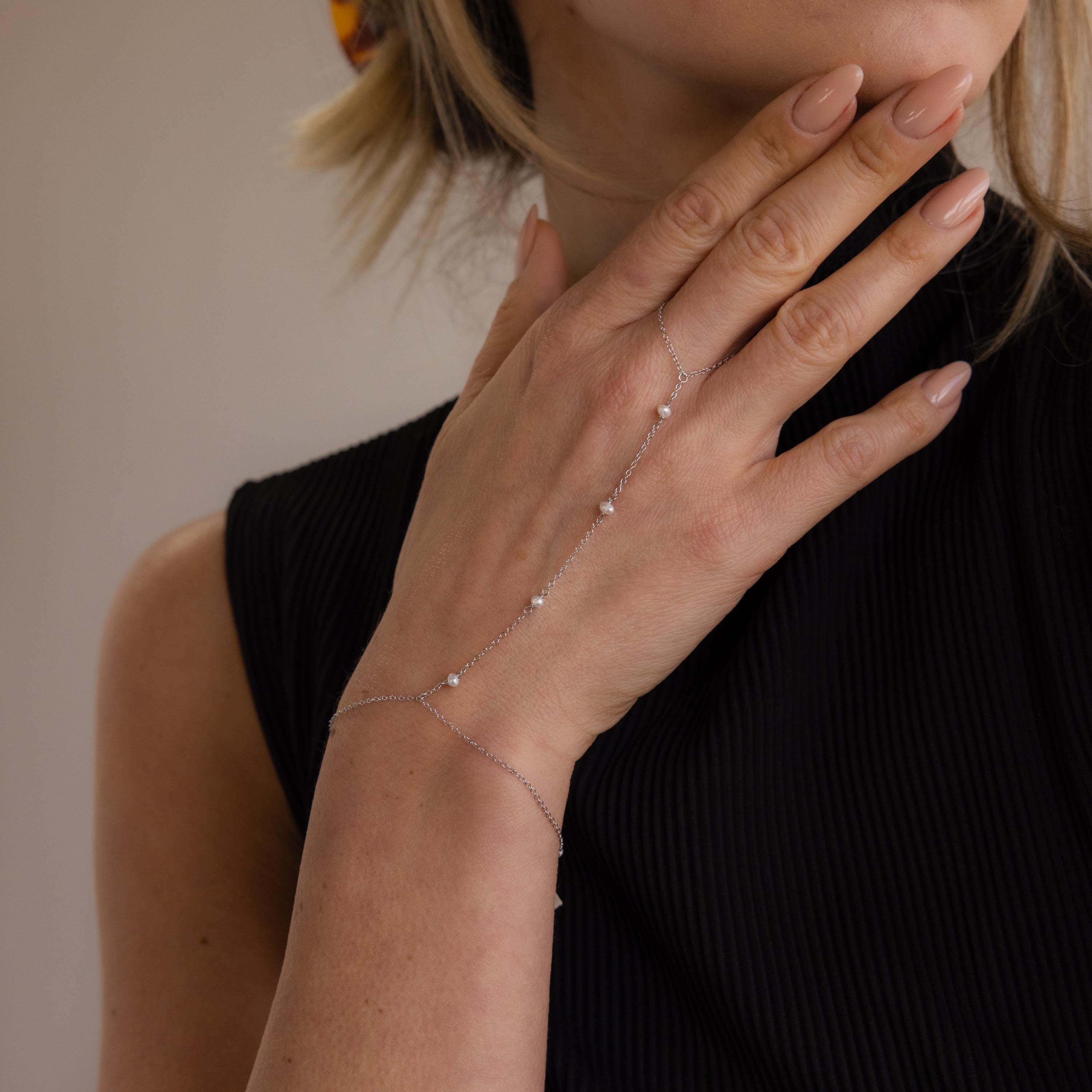A woman with nude nails wears the Pearl Hand Chain Bracelet, its small pearls adding summer elegance to her black sleeveless top.