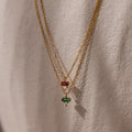Two gold chain necklaces with red and green gemstone pendants, including a Baguette Birthstone Drop Necklace, hanging against a textured beige background.