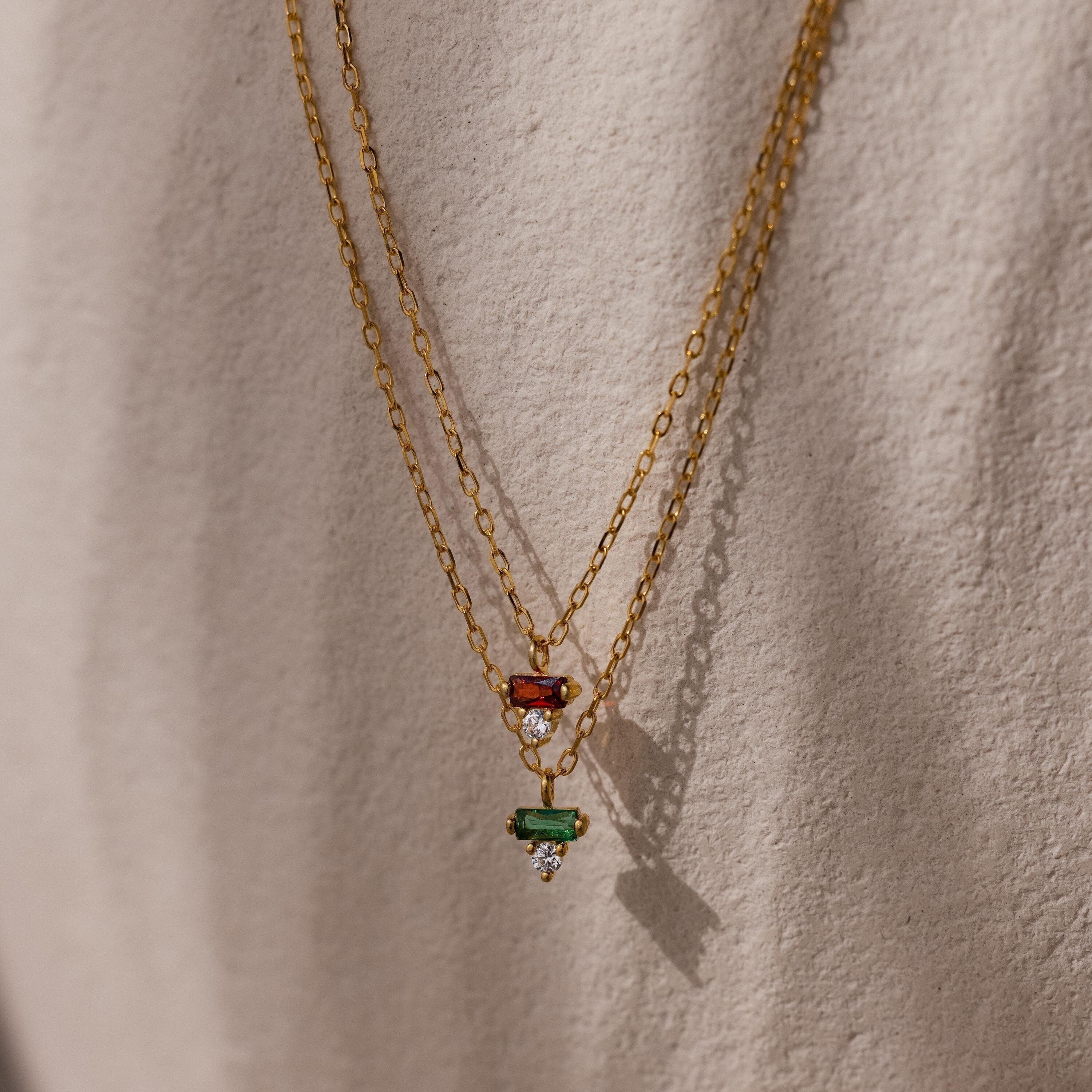 Two gold chain necklaces with red and green gemstone pendants, including a Baguette Birthstone Drop Necklace, hanging against a textured beige background.