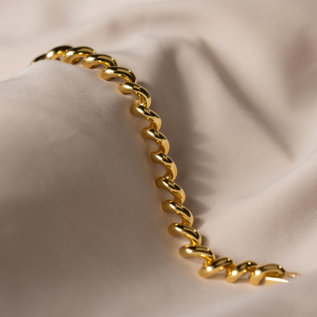 A Twisted Chain Bracelet with bold shining links rests on soft, light beige fabric with gentle folds, radiating effortless glamour.