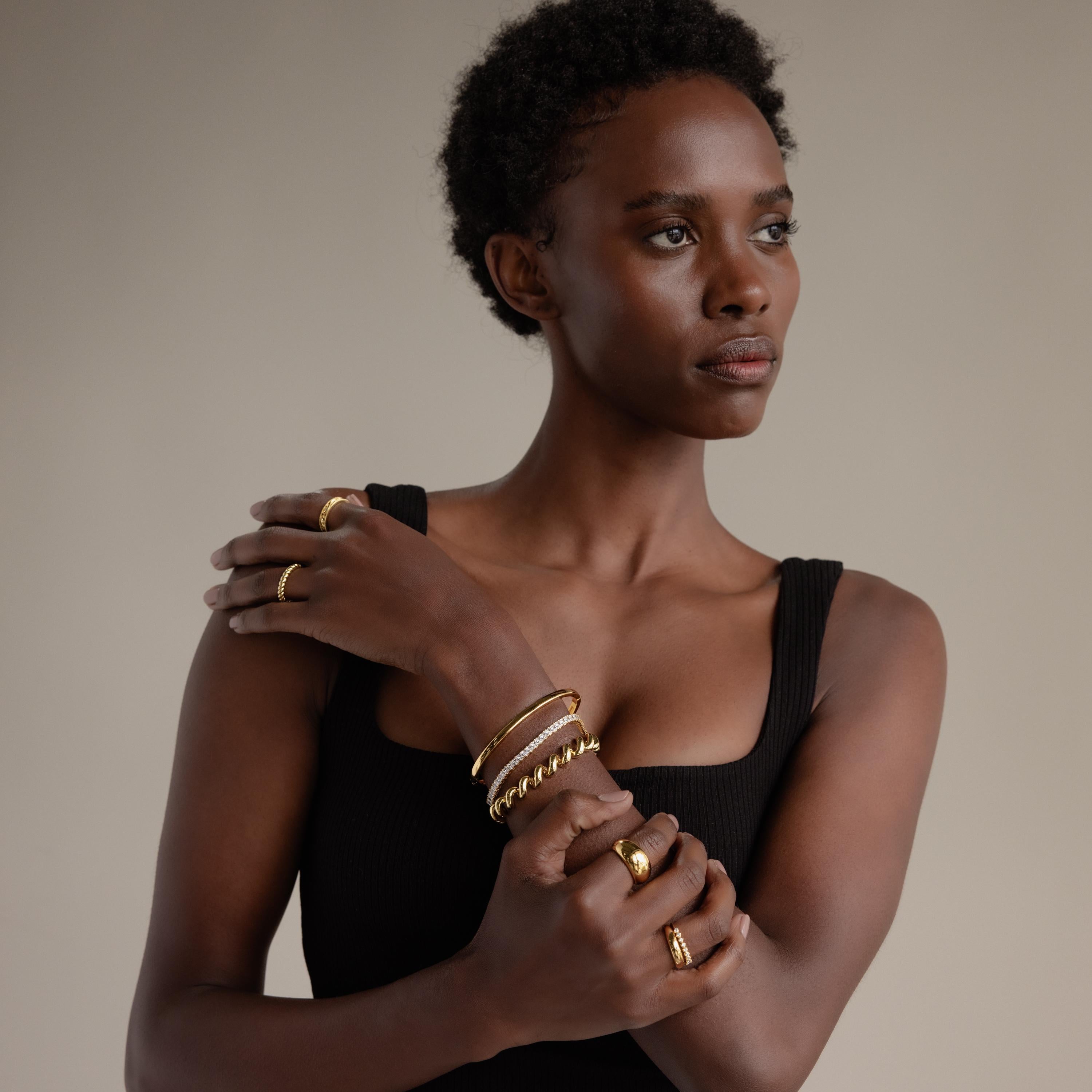 Woman in a black tank top, wearing bold shining links and multiple gold rings—including a Twisted Chain Bracelet—looks to the side, radiating effortless glamour against a neutral background.