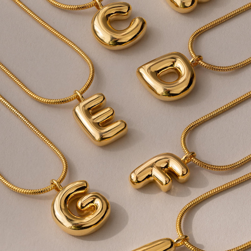 The Bubble Letter Necklace in 18K Gold features shiny bubble letter pendants—C, D, E, F, and G—arranged on a neutral background, ideal for anyone who loves personalized jewelry.