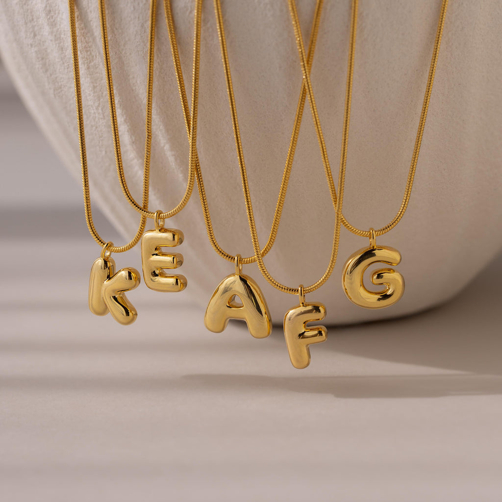 Four Bubble Letter Necklaces in 18K Gold—featuring chunky K, E, A, and G pendants—are displayed on a neutral background, adding a bold and playful touch to personalized jewelry.