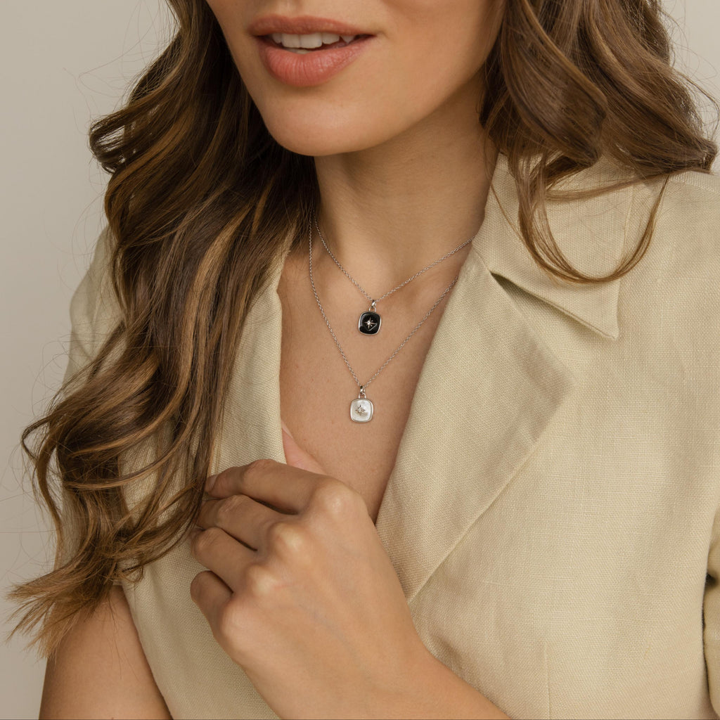 A woman in a beige blazer wears layered silver necklaces, including the striking Hermoine Pendant Necklace with contrasting black and white pendants, as she gently touches her collar.