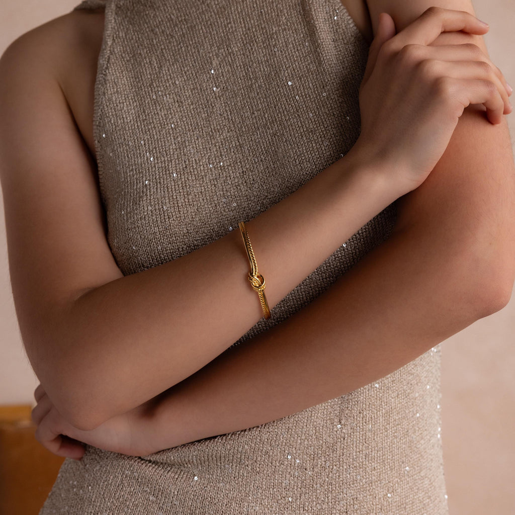 A woman in a sparkly beige dress with arms crossed wears the Duo Knot Bangle—an elegant gold bracelet featuring a rope texture and knot detail, making it a perfect gift.