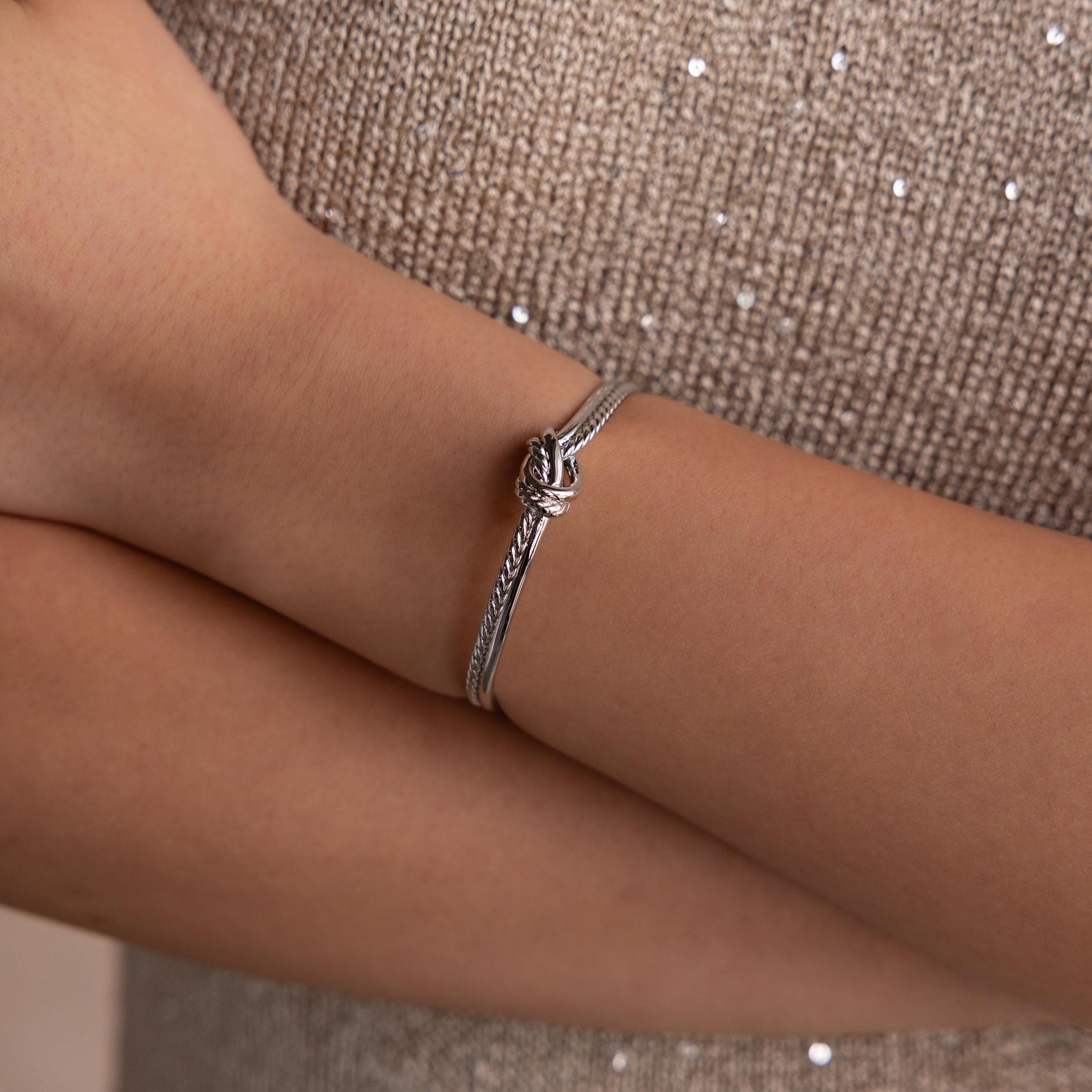 The Duo Knot Bangle, featuring a refined rope texture, adorns the wrist of someone in a sparkly beige top—an elegant gift choice for any occasion.
