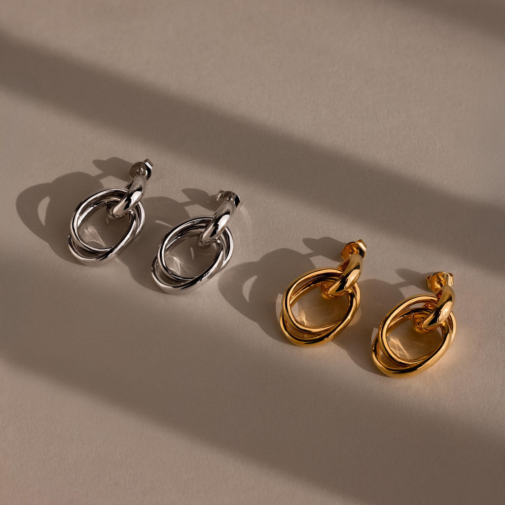 A pair of silver Bold Duo Knot Earrings and a pair of gold Bold Duo Knot Earrings lay on a beige surface, their sculptural, interlinked hoops casting elegant shadows.