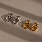 A pair of silver Bold Duo Knot Earrings and a pair of gold Bold Duo Knot Earrings lay on a beige surface, their sculptural, interlinked hoops casting elegant shadows.