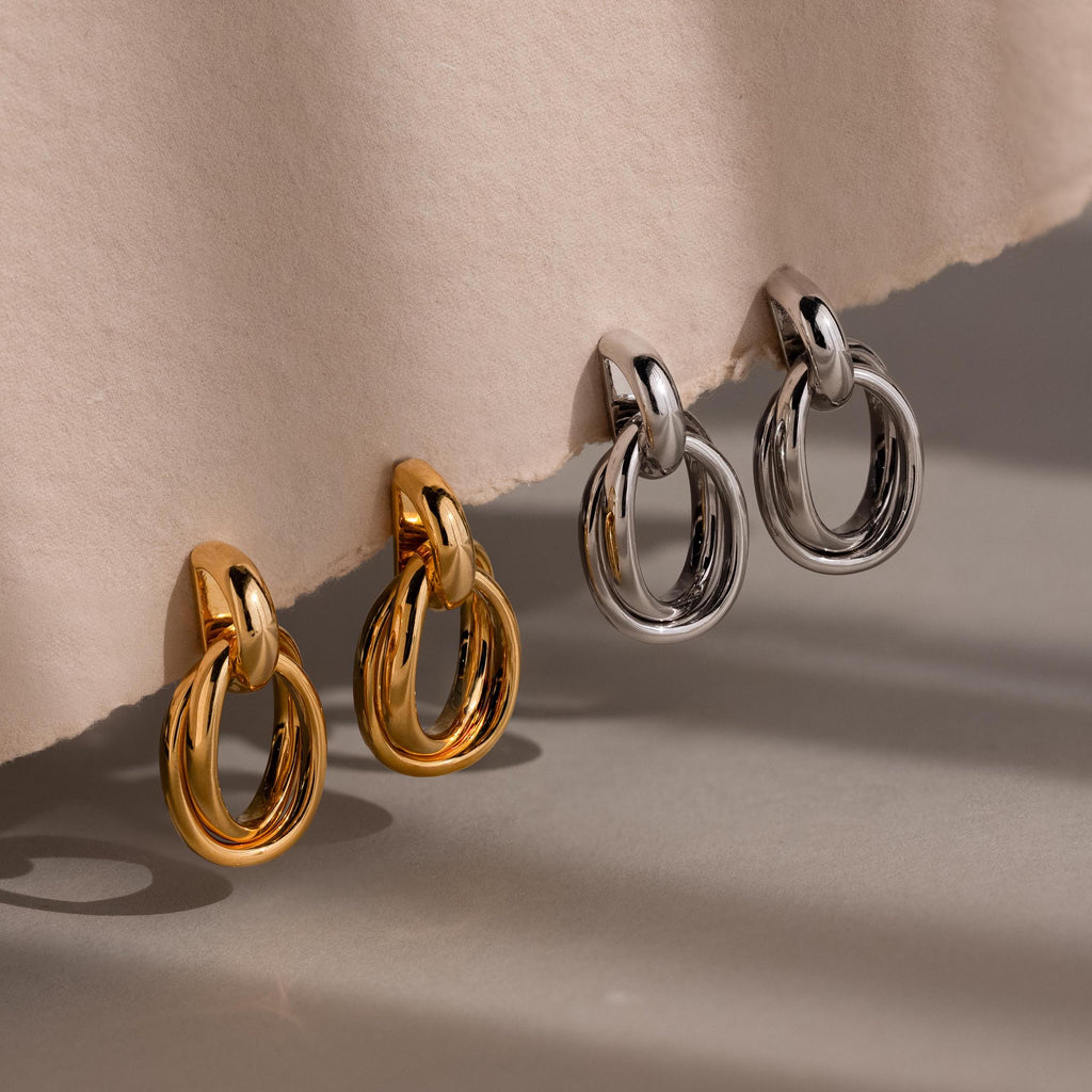 Two pairs of Bold Duo Knot Earrings, in gold and silver, rest on a beige textured surface. These sculptural earrings offer a modern update to your jewelry collection.