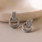 Ribbed Duo Knot Earrings rest on a smooth beige surface, showcasing their twisted rope design. The blurred background enhances these statement earrings, making them stand out beautifully with a modern touch.