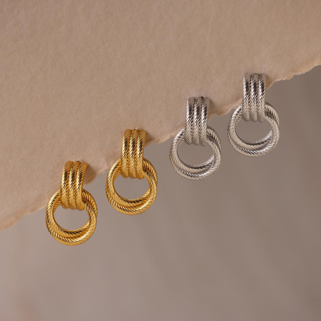 The Ribbed Duo Knot Earrings, featuring a textured hoop design in gold and silver, are displayed on a beige surface—perfect statement earrings for any occasion.