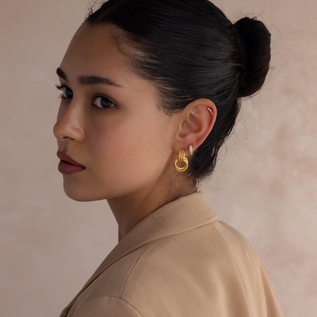 A woman with dark hair in a bun wears the Ribbed Duo Knot Earrings and a beige blazer, glancing over her shoulder.