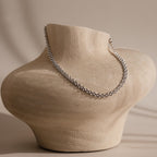 The Strap Necklace, a luxury silver chain with interwoven links, is displayed on a textured beige torso-shaped jewelry stand.