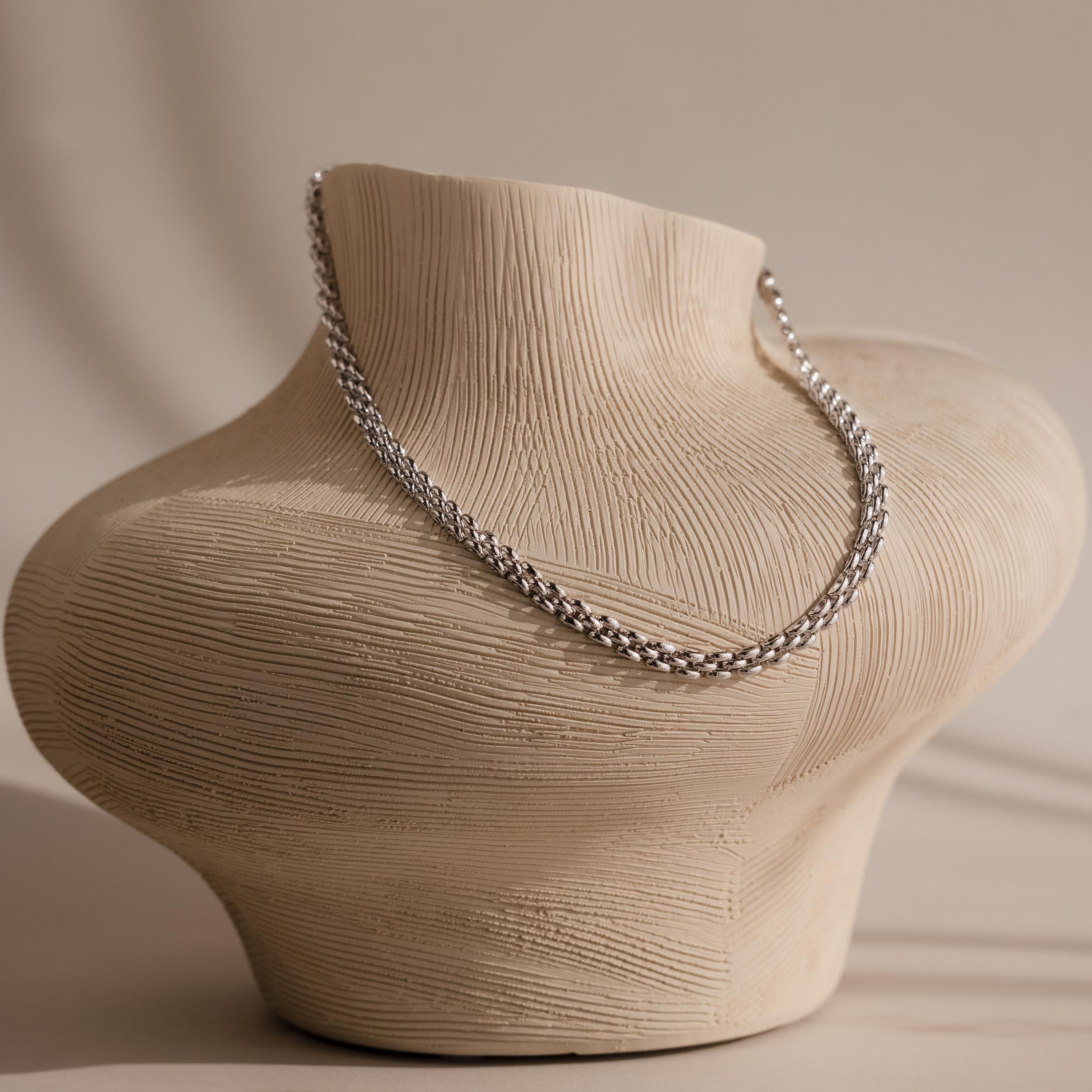 The Strap Necklace, a luxury silver chain with interwoven links, is displayed on a textured beige torso-shaped jewelry stand.