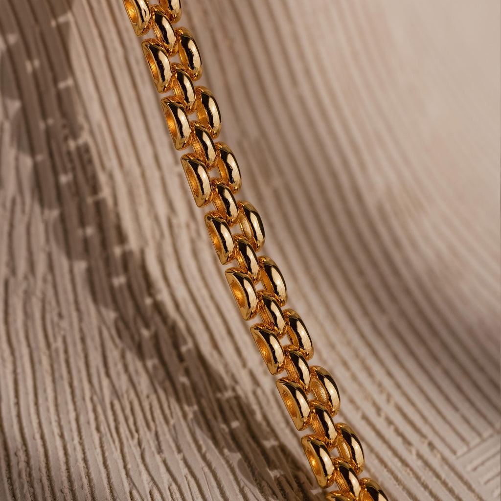 Close-up of the Strap Necklace, a luxury gold chain with interwoven links displayed on a textured beige background.