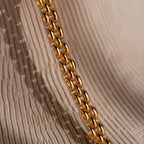 Close-up of the Strap Necklace, a luxury gold chain with interwoven links displayed on a textured beige background.