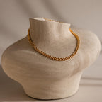 The Strap Necklace, a luxury gold chain with interwoven links, is displayed on a textured beige sculpted bust against a neutral background.