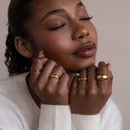 A woman with closed eyes softly poses with her hands near her face, wearing gold rings including the Mixed Birthstone Dome Ring, a customizable gemstone piece.