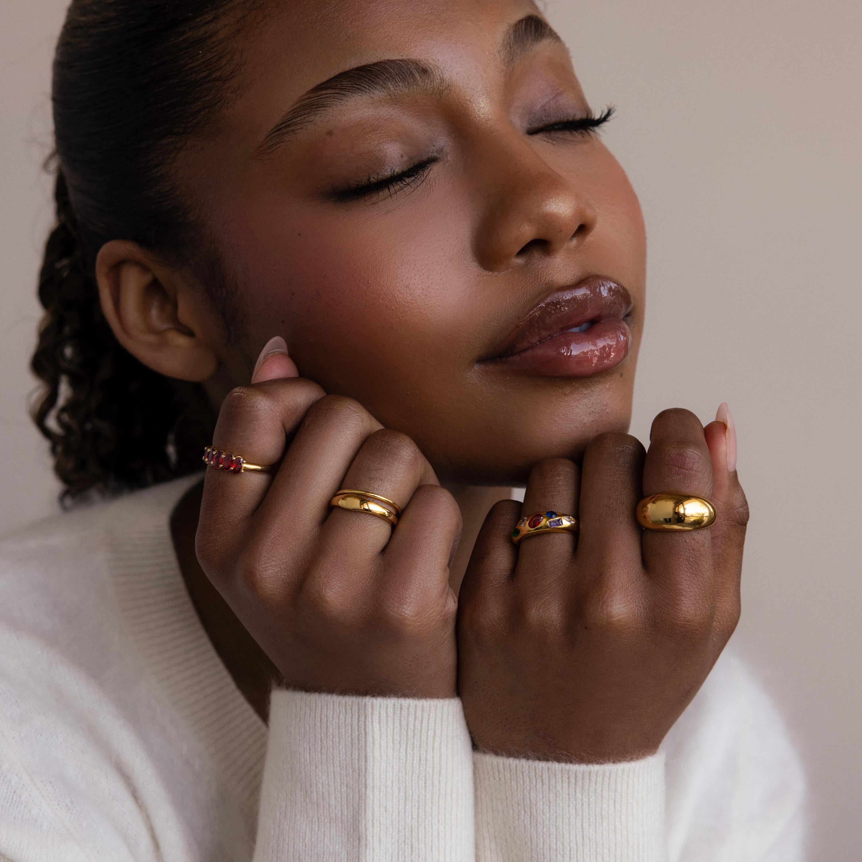 A woman with closed eyes softly poses with her hands near her face, wearing gold rings including the Mixed Birthstone Dome Ring, a customizable gemstone piece.