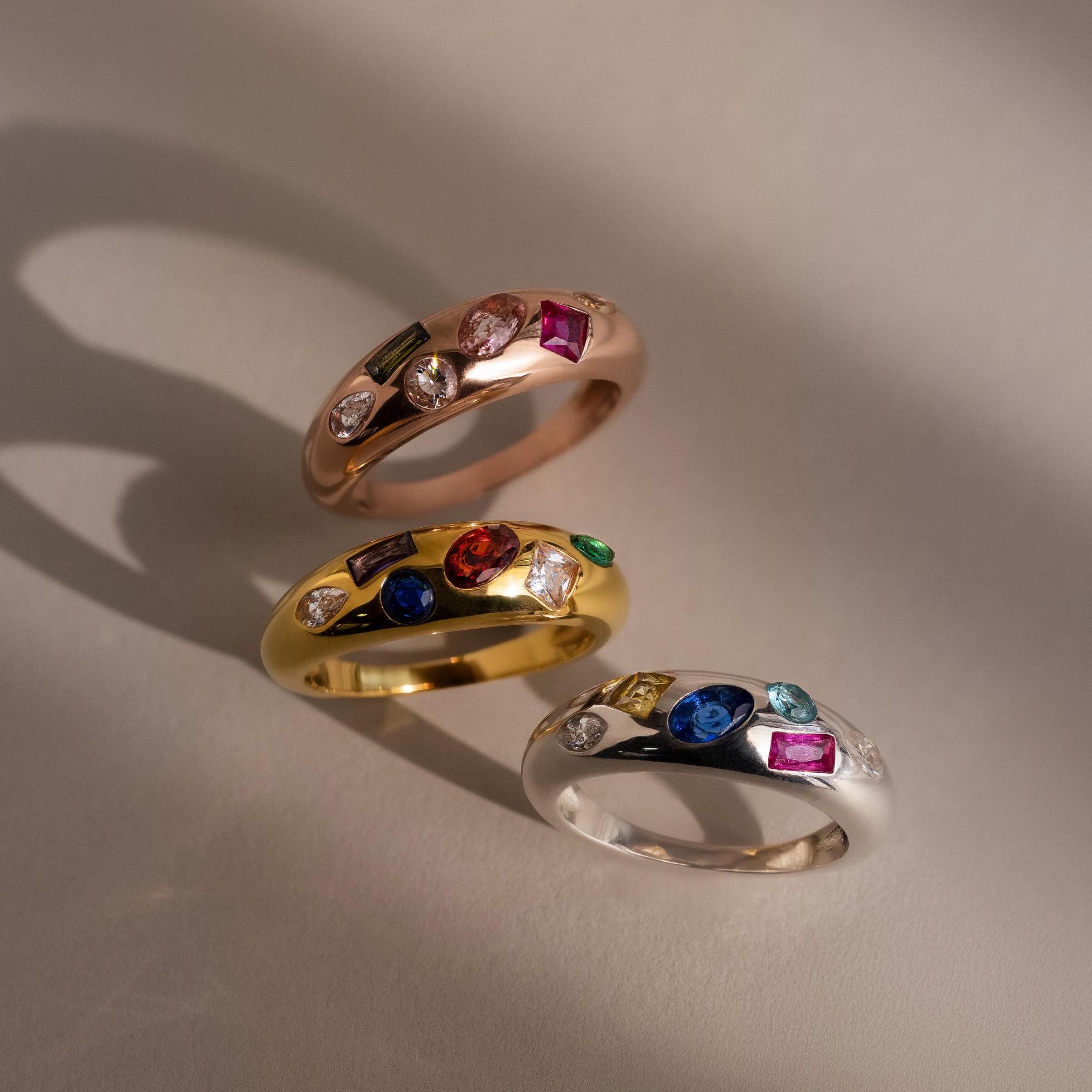 The Mixed Birthstone Dome Ring, available in gold, silver, and rose gold with vibrant gemstones and diamonds on a neutral background, is the perfect customizable jewelry piece for a personalized touch.
