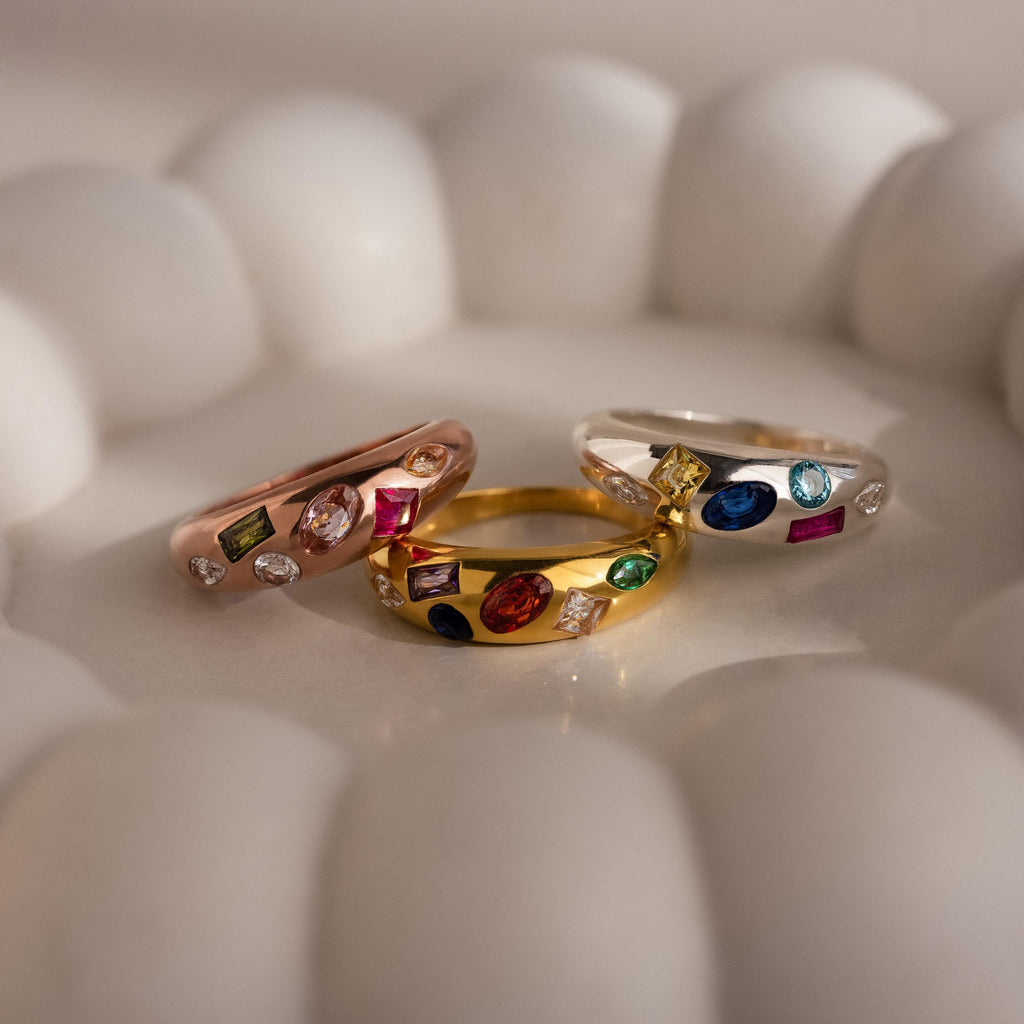 Three vibrant Mixed Birthstone Dome Rings in rose gold, yellow gold, and silver are displayed on a white scalloped dish—an ideal personalized jewelry piece for a unique touch.
