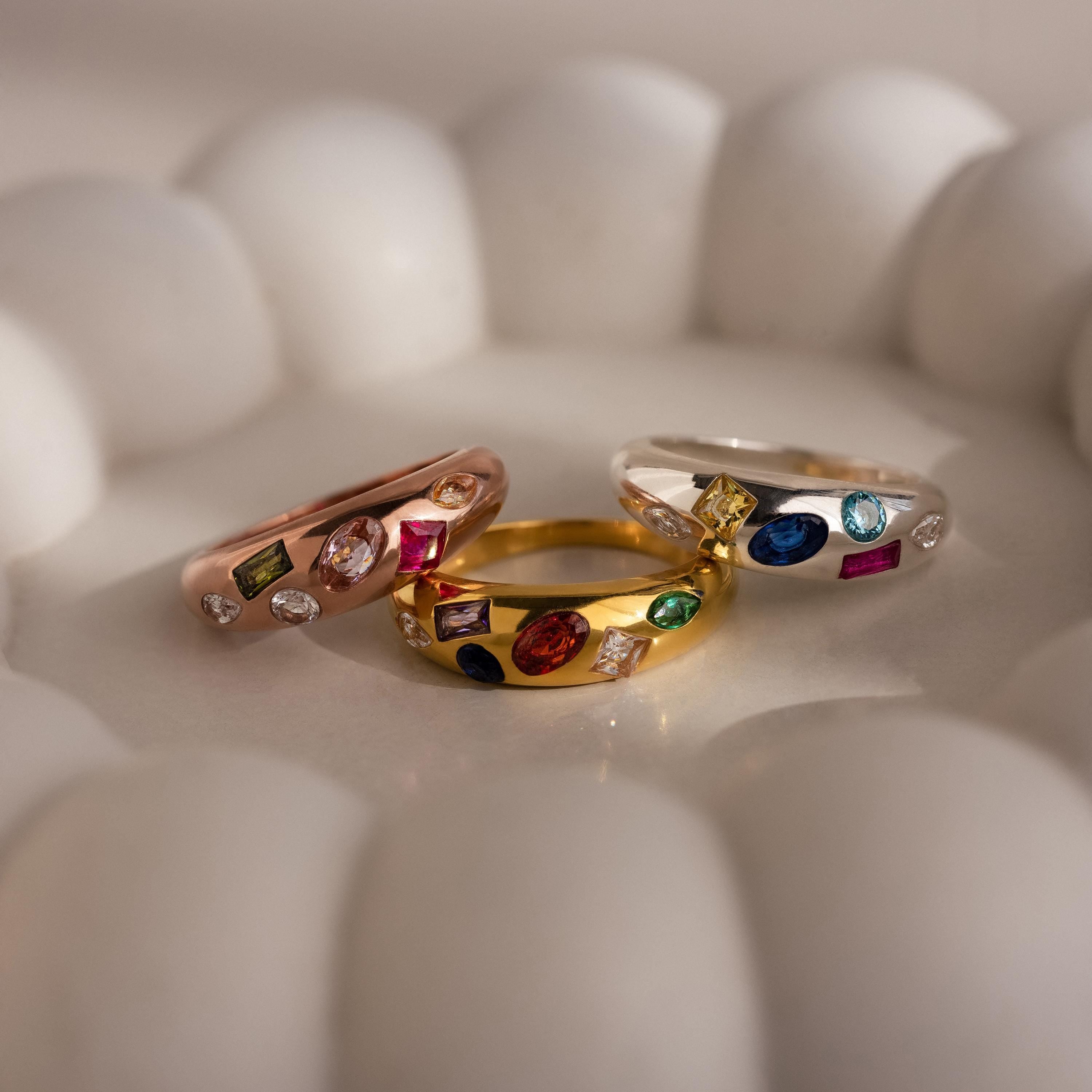 Three vibrant Mixed Birthstone Dome Rings in rose gold, yellow gold, and silver are displayed on a white scalloped dish—an ideal personalized jewelry piece for a unique touch.