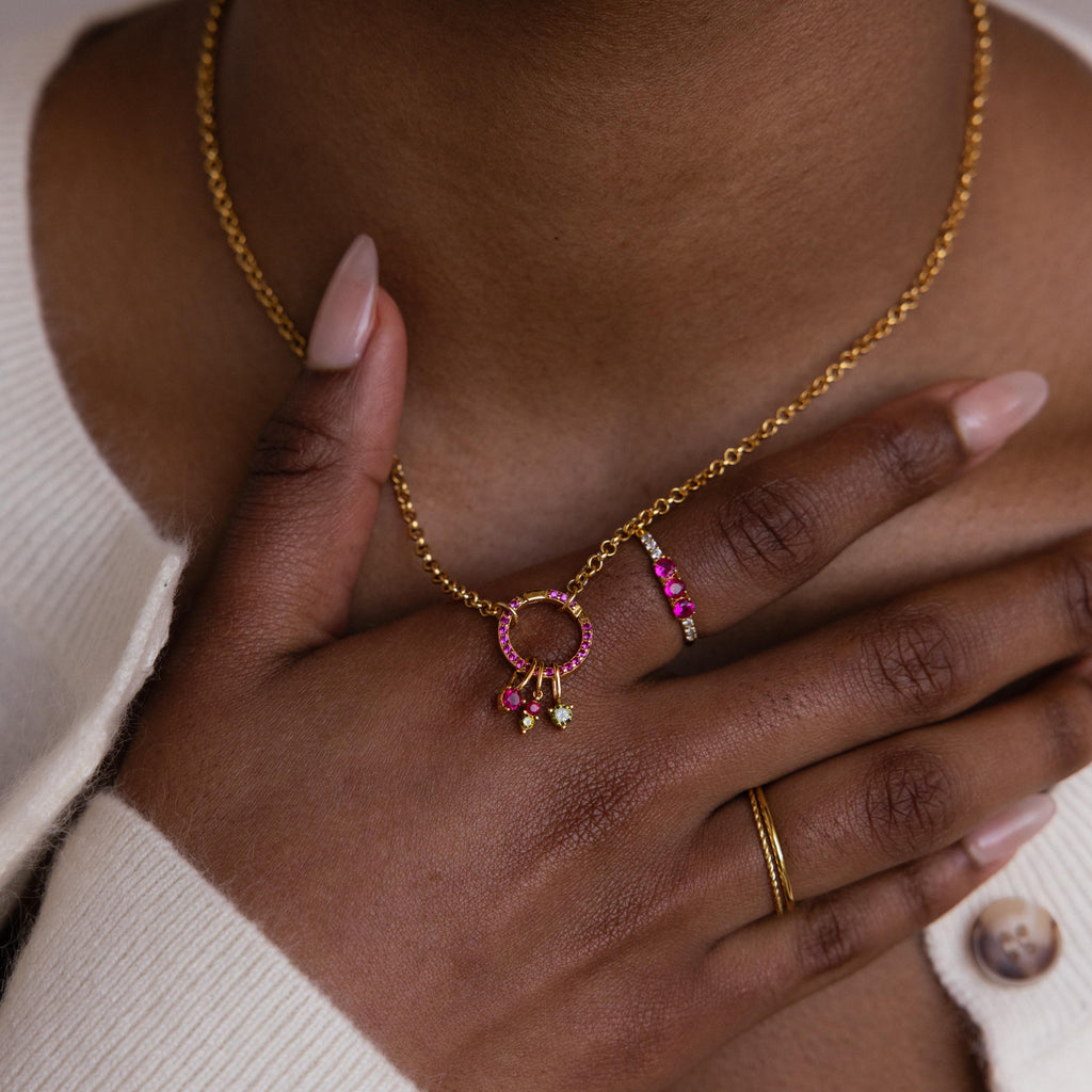 Model wears a gold chain with a pave birthstone clip. A Dainty Duo Birthstone Charm, and two other birthstone charm dangles off of the clip. Model completes the look with a matching ruby ring and duo stacking ring.