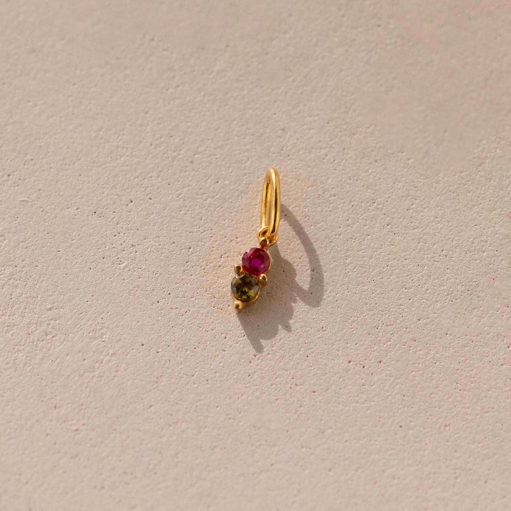 A gold Dainty Duo Birthstone Charm with ruby and peridot. The design features a ruby stone at the top and a peridot stone at the bottom held by prongs. The charm is attached to a large jump ring